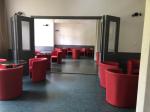 Widok sali kawiarnianej. Ściany w kolorze piaskowym. Sala składa się z dwóch części - na pierwszej widać czerwone fortele i stoły. Za nią rozsuwane przeszkolone drzwi i widok na kolejne fotele i stoliki.