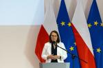 Minister finansów Magdalena Rzeczkowska stoi za mównicą, za jej plecami są flagi Polski i UE